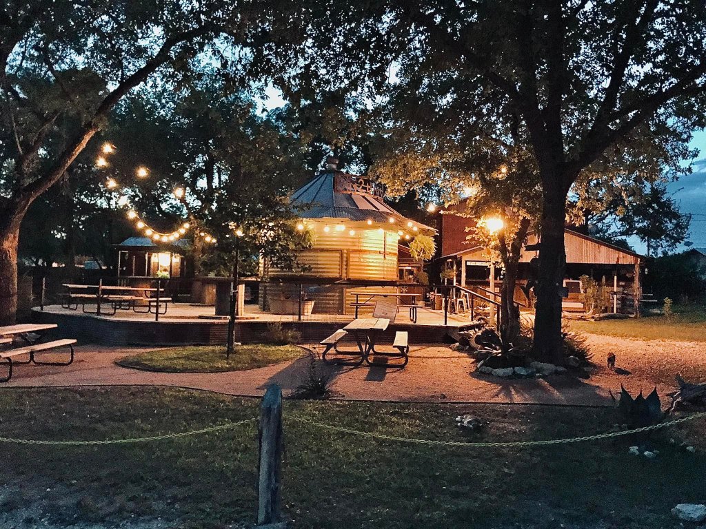 A serene outdoor dining area with string lights and wooden tables surrounded by lush trees at dusk in Uvalde County, Texas
