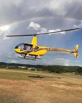 A vibrant yellow helicopter soars through the sky, its rotor blades spinning against a backdrop of a stunning rainbow arching over a lush green landscape in Uvalde County, Texas