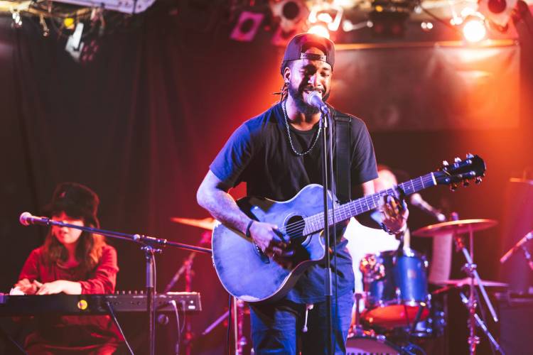 A musician performs on stage with an acoustic guitar, singing into a microphone, accompanied by a keyboardist and drummer in Uvalde County, Texas