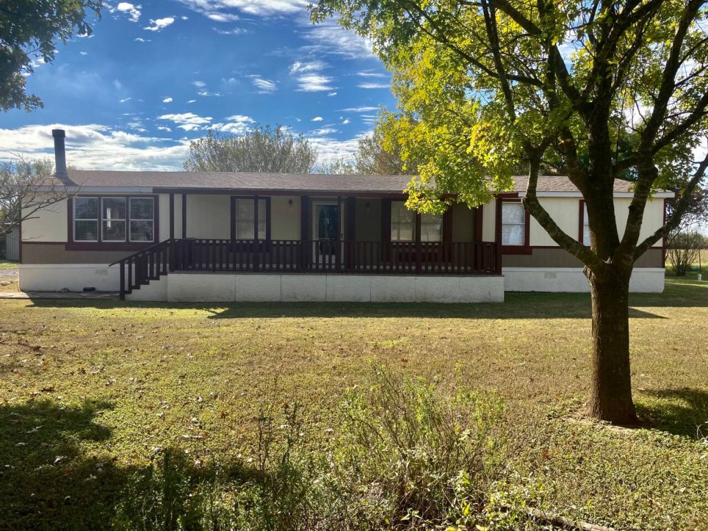 A single-story mobile home with a porch and a tree in the front yard, set against a backdrop of a clear blue sky with scattered clouds in Uvalde County, Texas