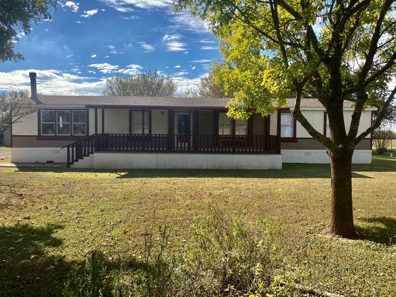 A single-story mobile home with a porch and a tree in the front yard, set against a backdrop of a clear blue sky with scattered clouds in Uvalde County, Texas