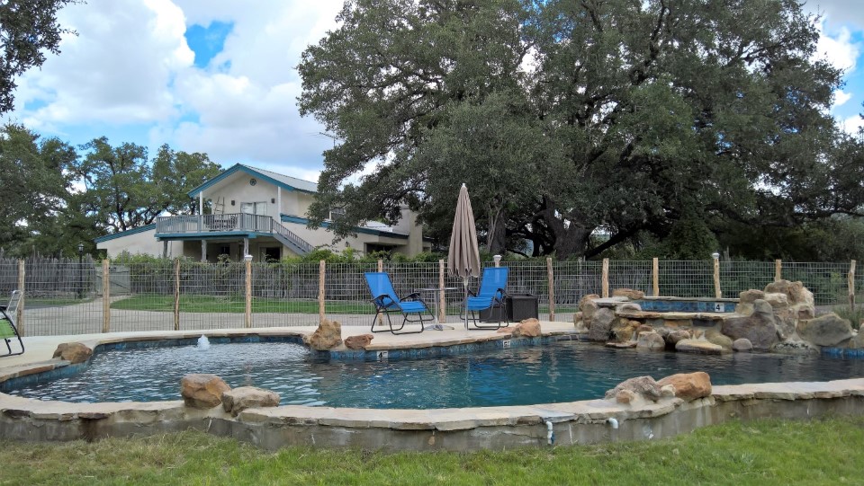 A serene backyard oasis featuring a natural stone swimming pool with a waterfall, surrounded by lush greenery and shaded by mature trees in Uvalde County, Texas