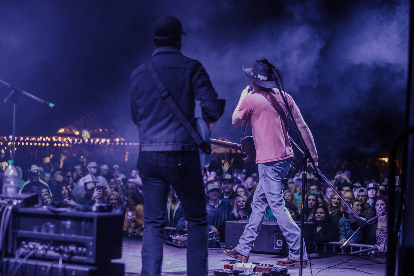 Two musicians perform on stage under a blue spotlight, with one playing a guitar and the other singing into a microphone, as a large crowd watches intently in Uvalde County, Texas