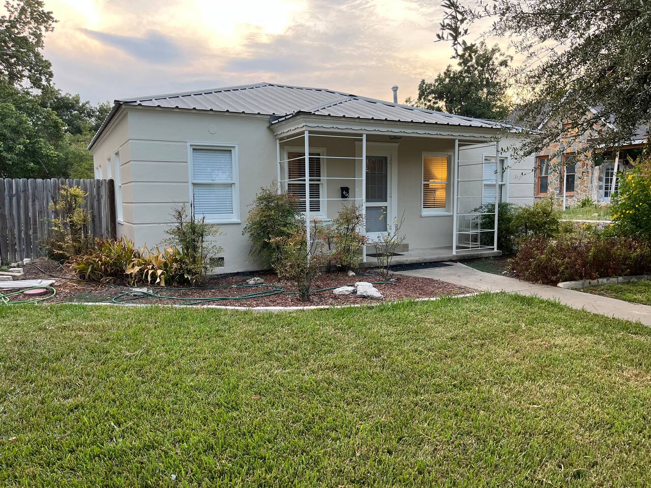 A quaint, single-story house with a white exterior and a covered front porch, nestled among lush greenery and a well-maintained lawn in Uvalde County, Texas