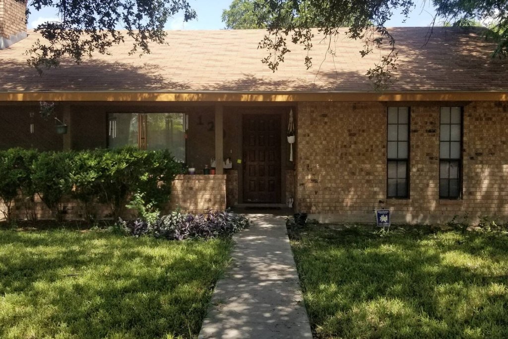 A single-story brick house with a covered front porch, surrounded by a well-maintained lawn and garden in Uvalde County, Texas