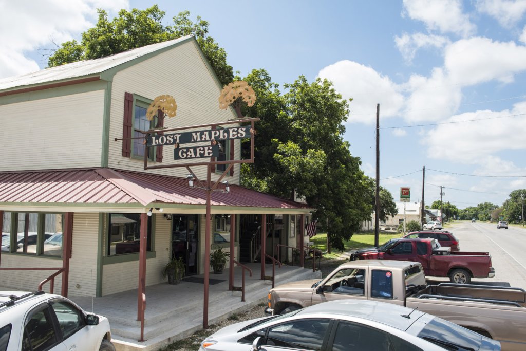 A quaint cafe with a rustic sign featuring maple leaves, nestled under a large tree in a small town setting in Uvalde County, Texas