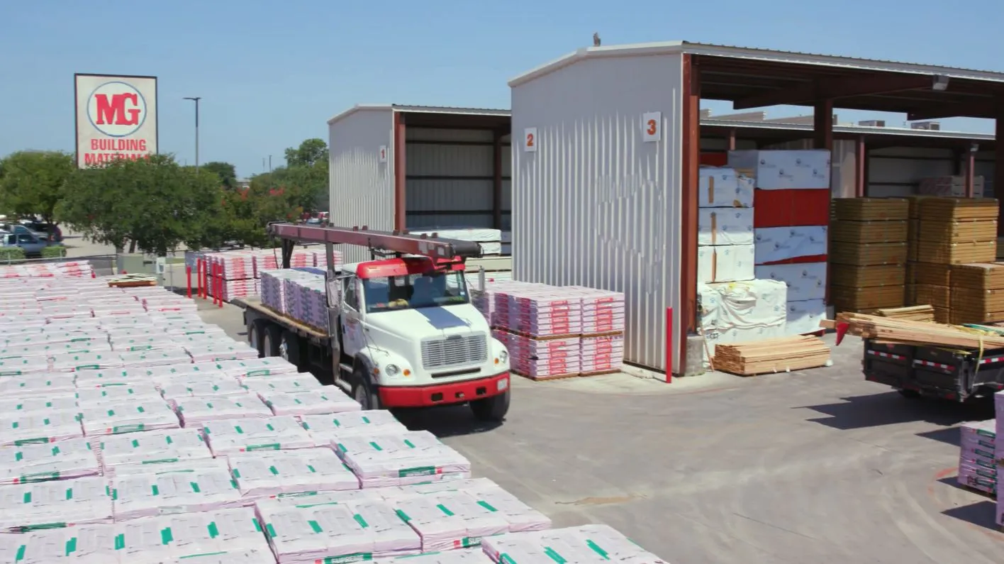 A large warehouse with a red and white MG sign, surrounded by stacks of building materials and a red truck with a crane in Uvalde County, Texas