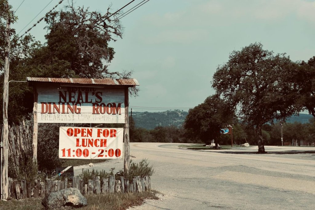 A rustic sign for Neal's Dining Room stands by the side of a quiet road, inviting passersby for lunch from 11:00 to 2:00 in Uvalde County, Texas