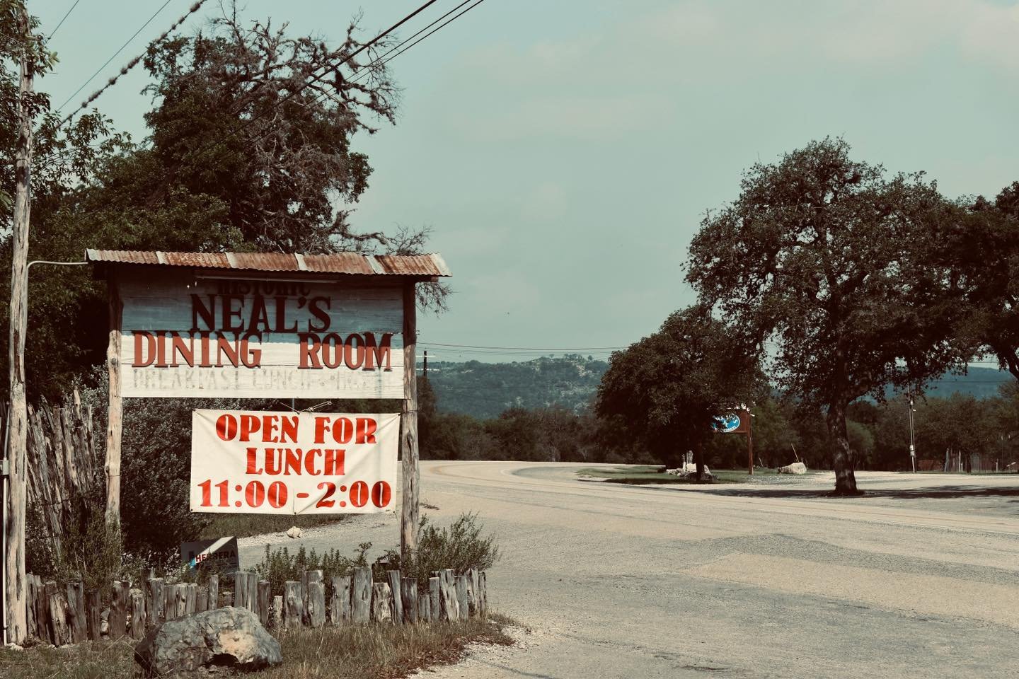 A rustic sign for Neal's Dining Room stands by the side of a quiet road, inviting passersby for lunch from 11:00 to 2:00 in Uvalde County, Texas