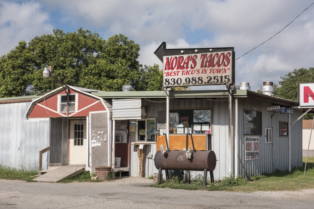 A rustic taco stand with a red and white exterior, featuring a large sign that reads 'Nora's Tacos' and a phone number in Uvalde County, Texas
