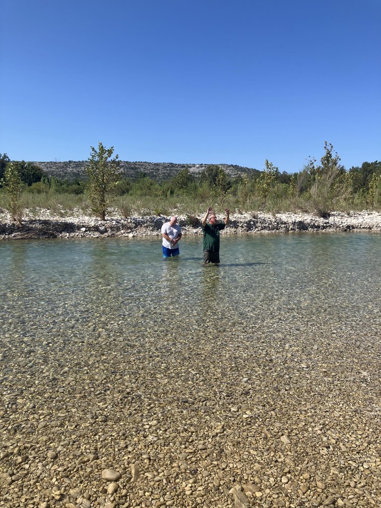 Two people are standing in a shallow, clear river with pebbles on the bottom, surrounded by a natural landscape with trees and hills in the background in Uvalde County, Texas
