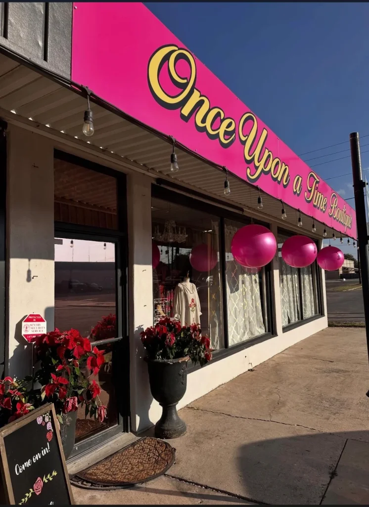 A vibrant pink sign with yellow lettering reading 'Once Upon a Time Boutique' hangs above a charming storefront decorated with red poinsettias and pink balloons in Uvalde County, Texas