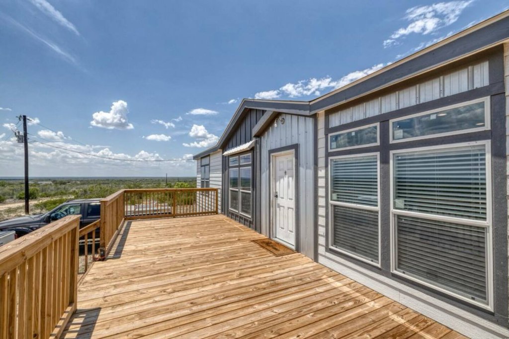 A spacious wooden deck extends from a modern, gray and white manufactured home, offering a panoramic view of the surrounding Texas landscape under a clear blue sky in Uvalde County, Texas