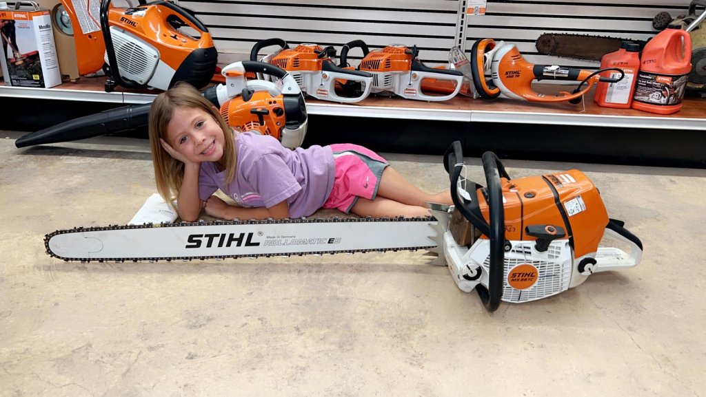 A young girl is lying on the floor of a hardware store, surrounded by various chainsaws and outdoor power equipment, smiling as she poses with a large STIHL chainsaw beside her in Uvalde County, Texas