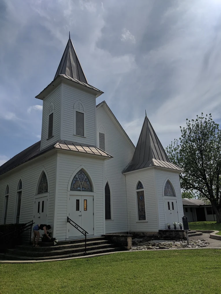 A quaint white church with two steepled towers and arched windows stands in a serene grassy area, with a few people gathered near the entrance in Uvalde County, Texas