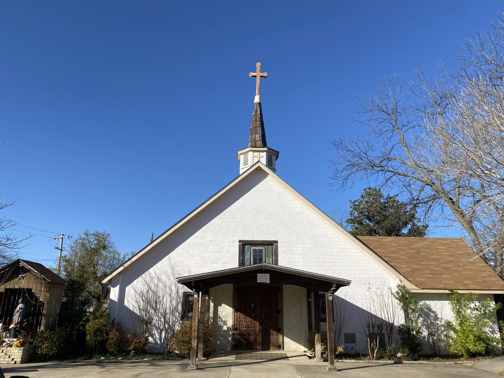 A quaint white church with a tall steeple and cross stands under a clear blue sky, surrounded by lush greenery and a serene atmosphere in Uvalde County, Texas