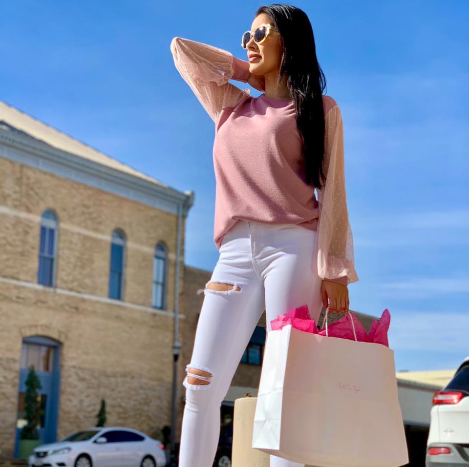 A woman in a pink top and white jeans holds shopping bags outside a brick building on a sunny day in Uvalde County, Texas