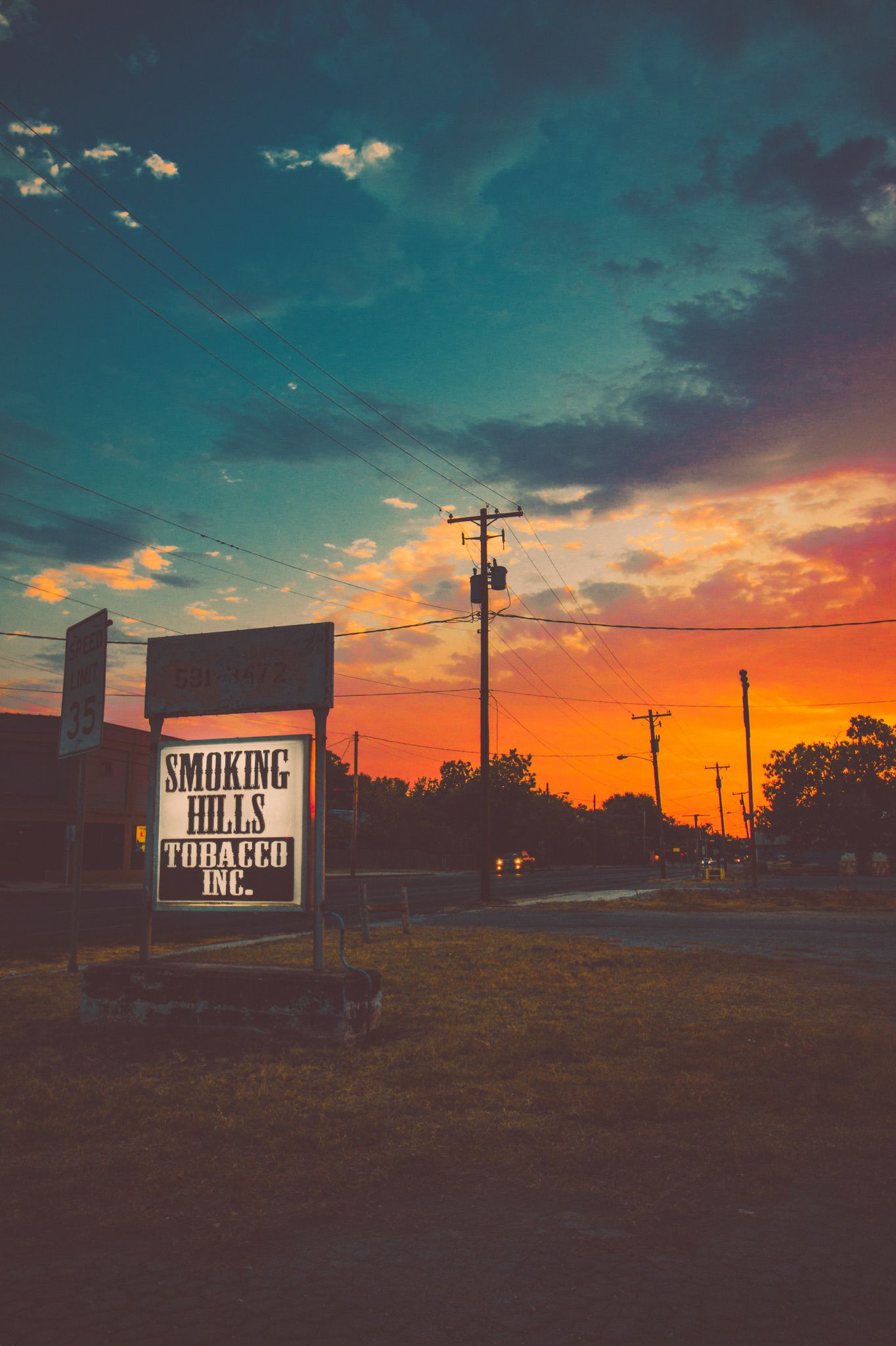 A vibrant sunset paints the sky with hues of orange and blue, casting a warm glow over a rural landscape featuring a sign for Smoking Hills Tobacco Inc. and power lines stretching into the distance in Uvalde County, Texas