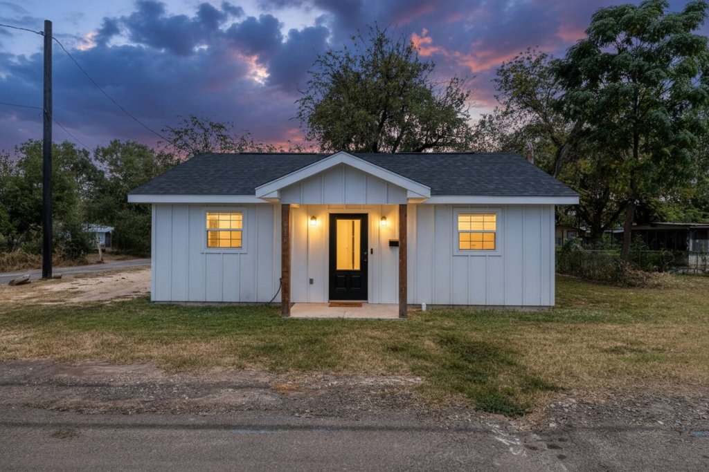 A quaint, white, one-story house with a dark roof and wooden accents sits on a grassy lot at dusk, with warm lights glowing from its windows and door in Uvalde County, Texas