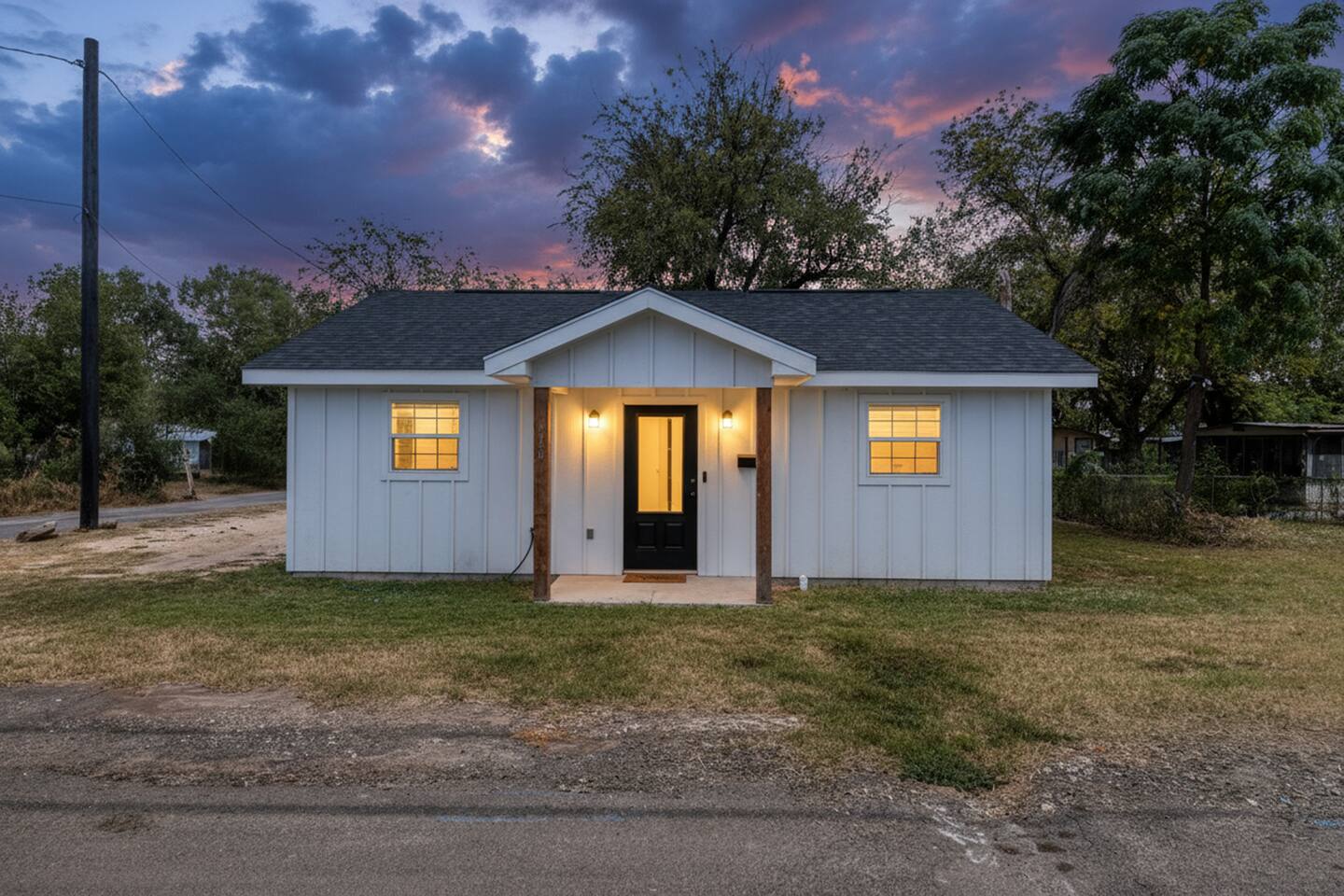A quaint, white, one-story house with a dark roof and wooden accents sits on a grassy lot at dusk, with warm lights glowing from its windows and door in Uvalde County, Texas