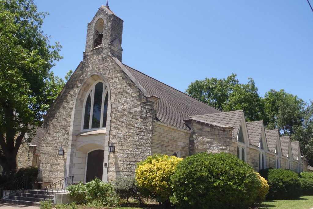 A quaint stone church with a tall bell tower and arched windows, nestled among lush greenery and blooming yellow flowers in Uvalde County, Texas