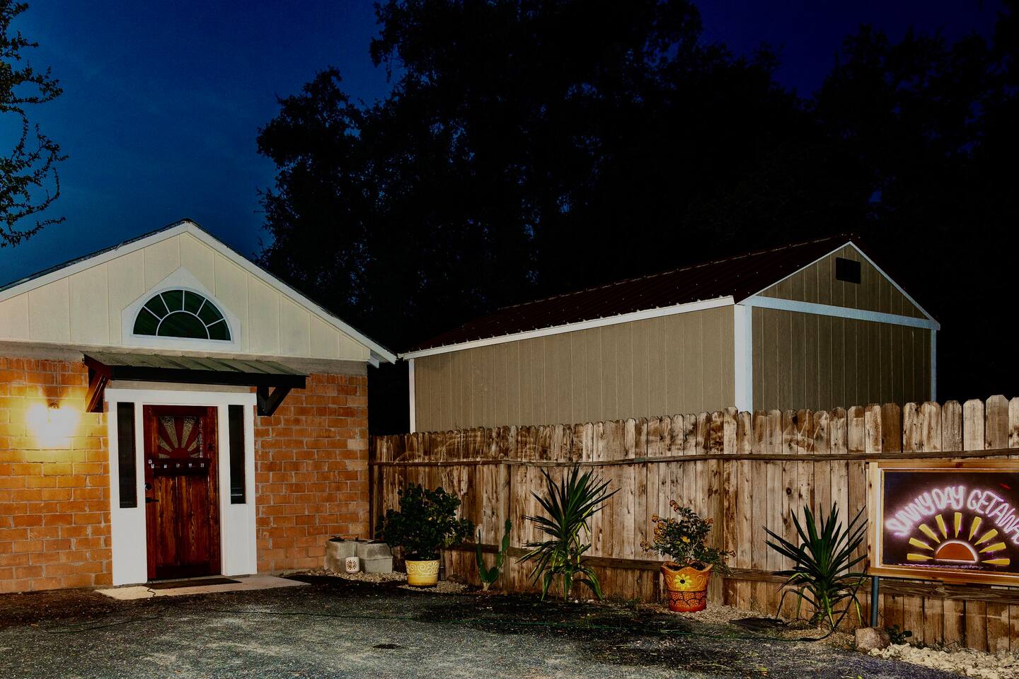 A quaint, single-story building with a white-framed arched window and a wooden door stands next to a larger, beige shed, both enclosed by a wooden fence with potted plants along the perimeter in Uvalde County, Texas