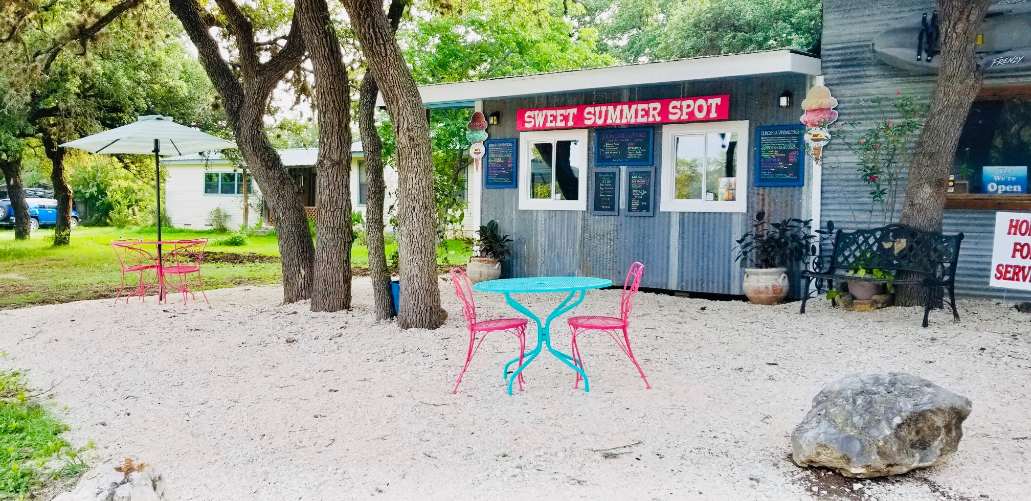 A quaint outdoor cafe named Sweet Summer Spot with a colorful turquoise table and pink chairs under a tree, surrounded by a sandy area and lush greenery in Uvalde County, Texas