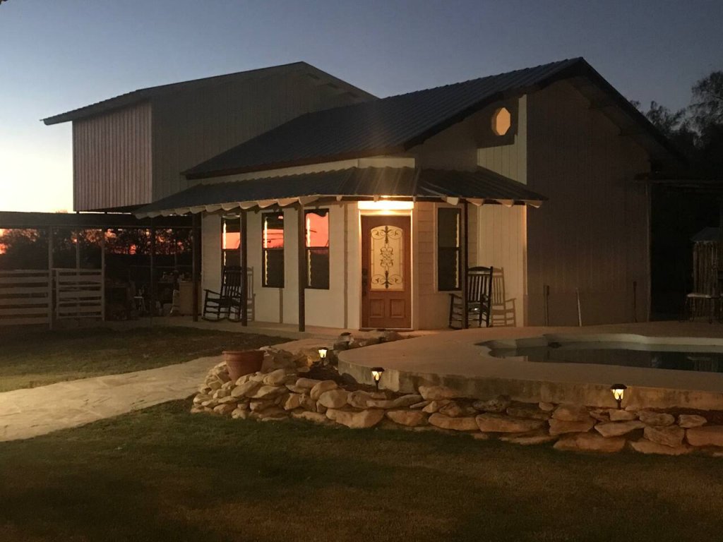 A rustic, two-story house with a wrap-around porch and a swimming pool, illuminated by soft outdoor lighting at dusk in Uvalde County, Texas