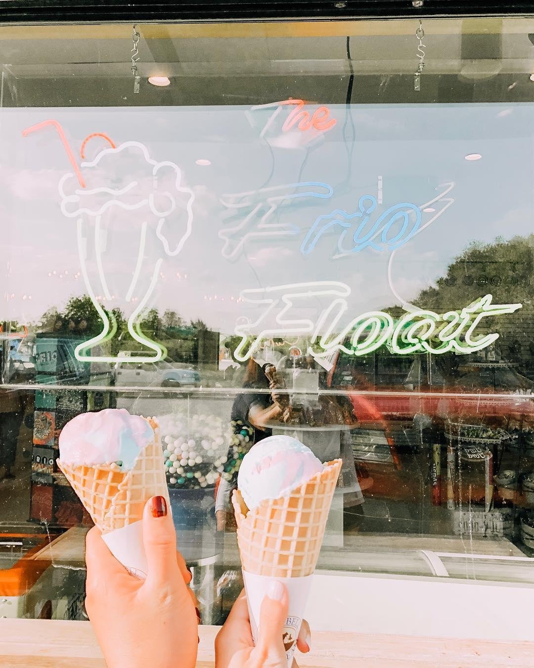 Two hands holding ice cream cones with pink and blue swirled ice cream in front of a neon sign that reads 'Frio Float' in Uvalde County, Texas