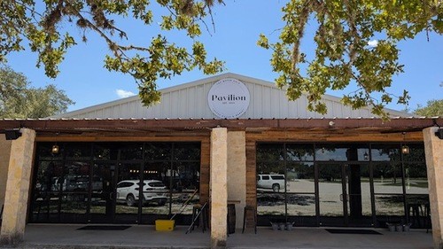 A modern, open-concept building with large glass windows and a wooden facade, surrounded by lush greenery and a clear blue sky in Uvalde County, Texas