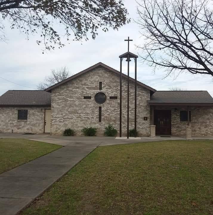 A simple, modern church building with a cross and bell tower, set in a grassy area with a paved pathway in Uvalde County, Texas