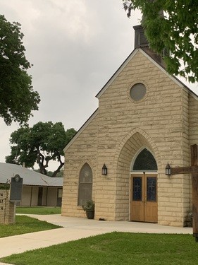A quaint stone church with a pointed arch entrance and a small bell tower stands amidst lush greenery and mature trees in Uvalde County, Texas