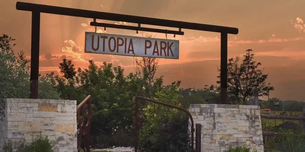 A rustic sign for Utopia Park stands against a backdrop of a vibrant sunset, with trees and a stone wall framing the entrance in Uvalde County, Texas