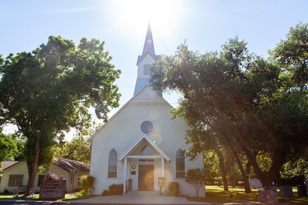 A quaint white church with a tall steeple stands amidst lush green trees, bathed in the warm glow of the setting sun in Uvalde County, Texas