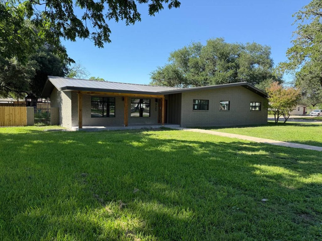A modern single-story house with a flat roof and large windows is nestled among lush green trees on a sunny day in Uvalde County, Texas