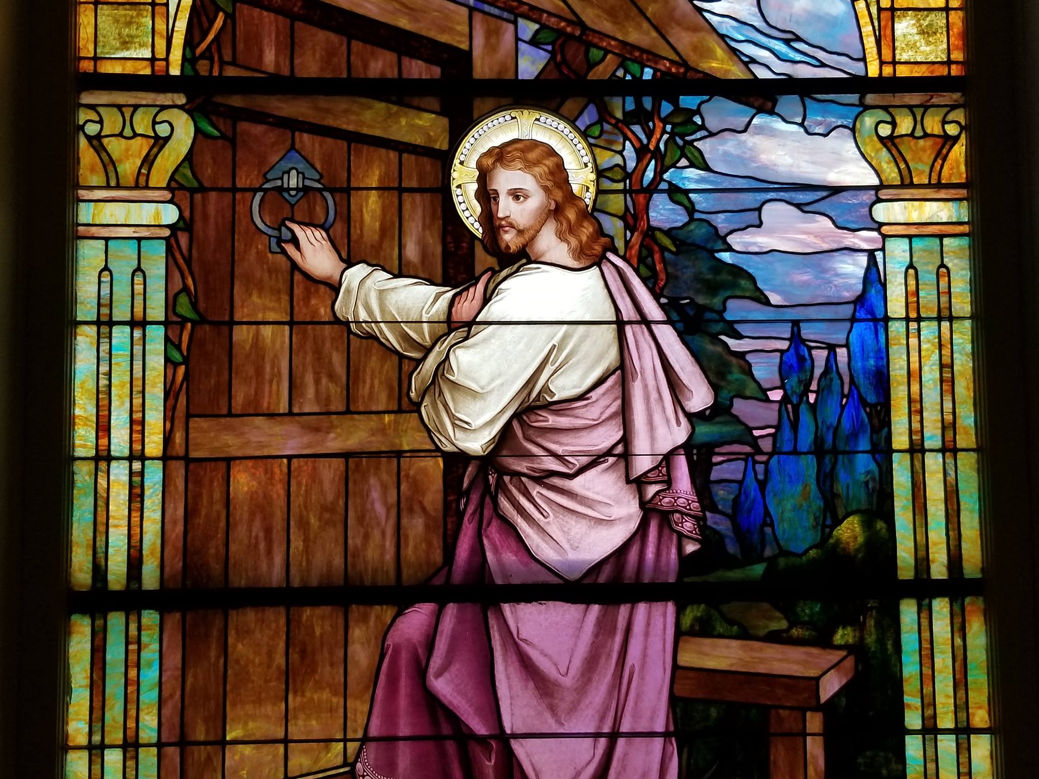 Stained glass window depicting Jesus knocking on a door, surrounded by lush greenery and a serene sky in Uvalde County, Texas