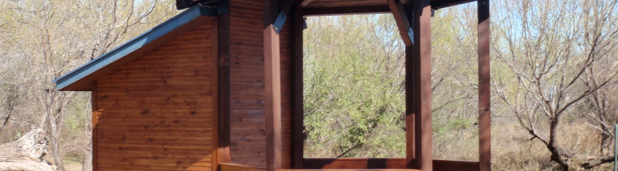 A rustic wooden structure with a slanted roof and large glass windows, nestled among trees in a natural setting in Uvalde County, Texas