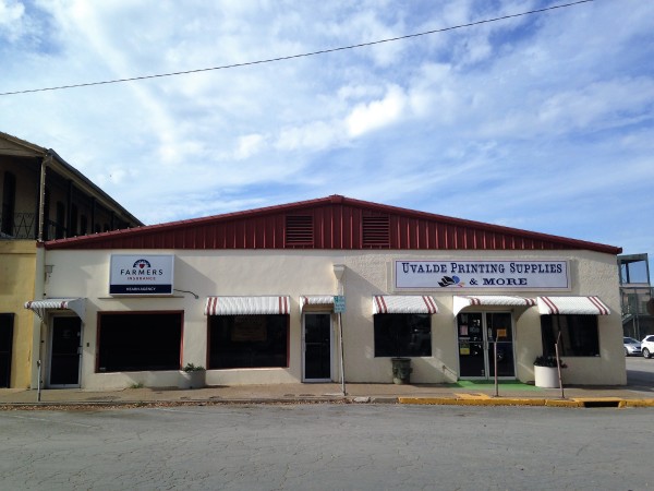 A commercial building with a red roof and white walls houses Uvalde Printing Supplies & More and Farmers Insurance in Uvalde County, Texas