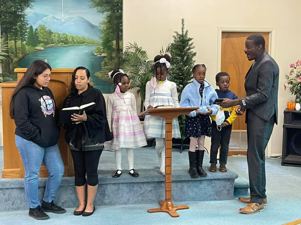 A group of children and adults stand on a stage in a church, with a man in a suit addressing them from a podium in Uvalde County, Texas