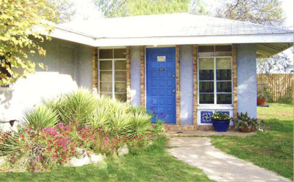 A quaint, single-story house with a bright blue door and matching window frames, nestled among lush greenery and vibrant flowers in a well-maintained front yard in Uvalde County, Texas