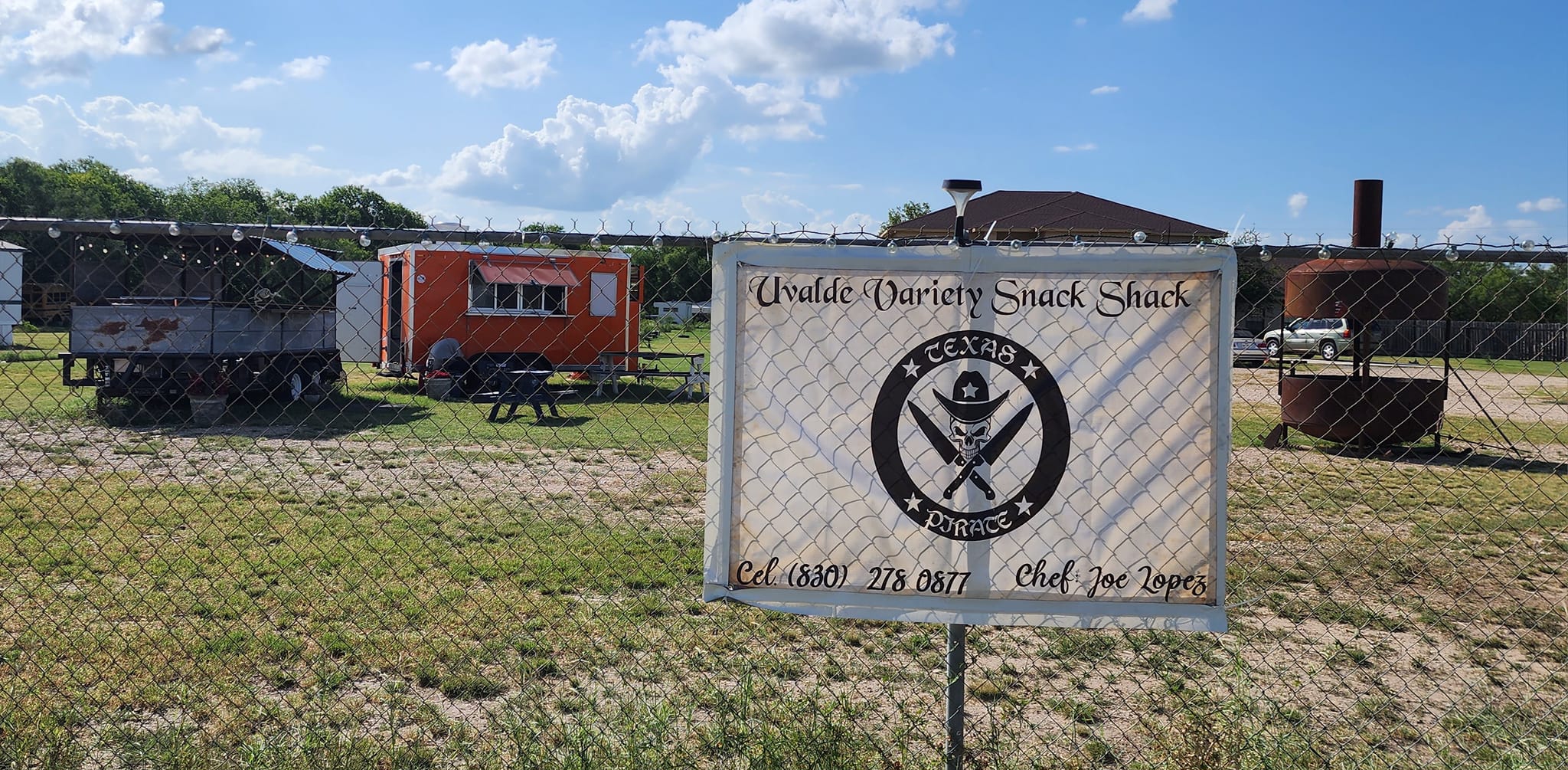 A sign for 'Wilde Oak Snack Shack' with a skull and crossbones logo stands in a grassy field next to food trucks and a fenced area in Uvalde County, Texas