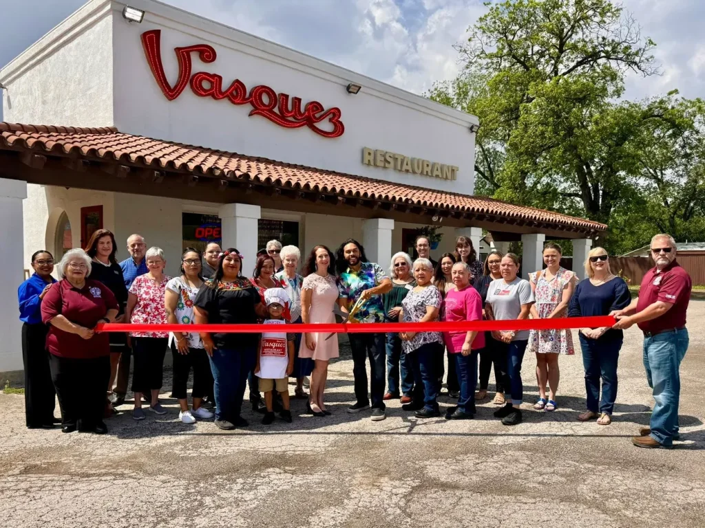 A diverse group of people stands in front of a restaurant named 'Vasques' for a ribbon-cutting ceremony, celebrating a grand opening or special event in Uvalde County, Texas