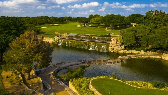 Aerial view of a serene golf course with a waterfall and pond surrounded by lush greenery and a stone bridge in Uvalde County, Texas