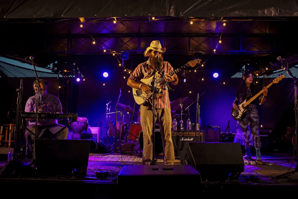 An image of a man in a cowboy hat playing guitar on a stage with blue lights. there is a woman to his right playing bass, a man to his left playing the keyboard, and a drumkit behind him