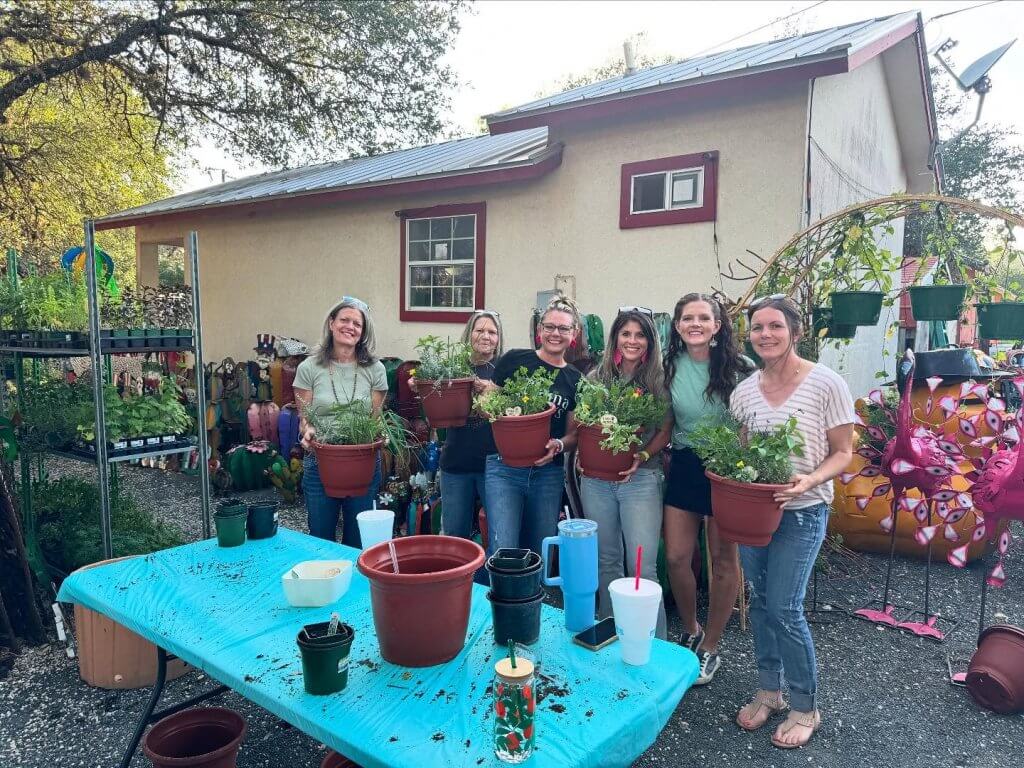 Six women smiling and holding potted plants outdoors at Concan Nursery.