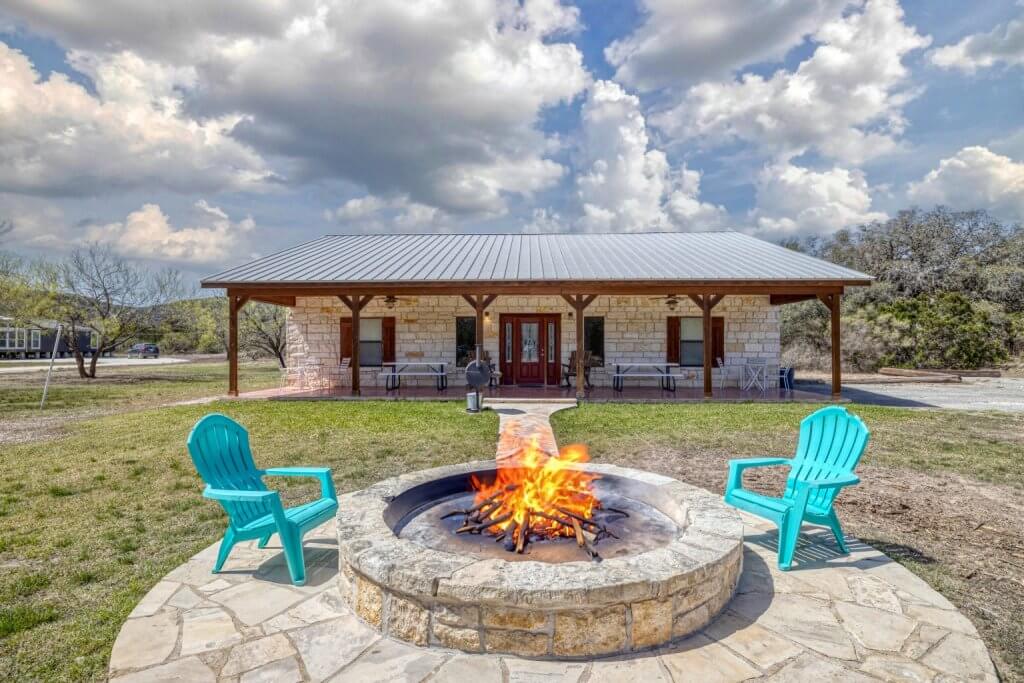 An image of a 1 story stone cabin with a porch, two blue chairs are in front of the cabin by a firepit