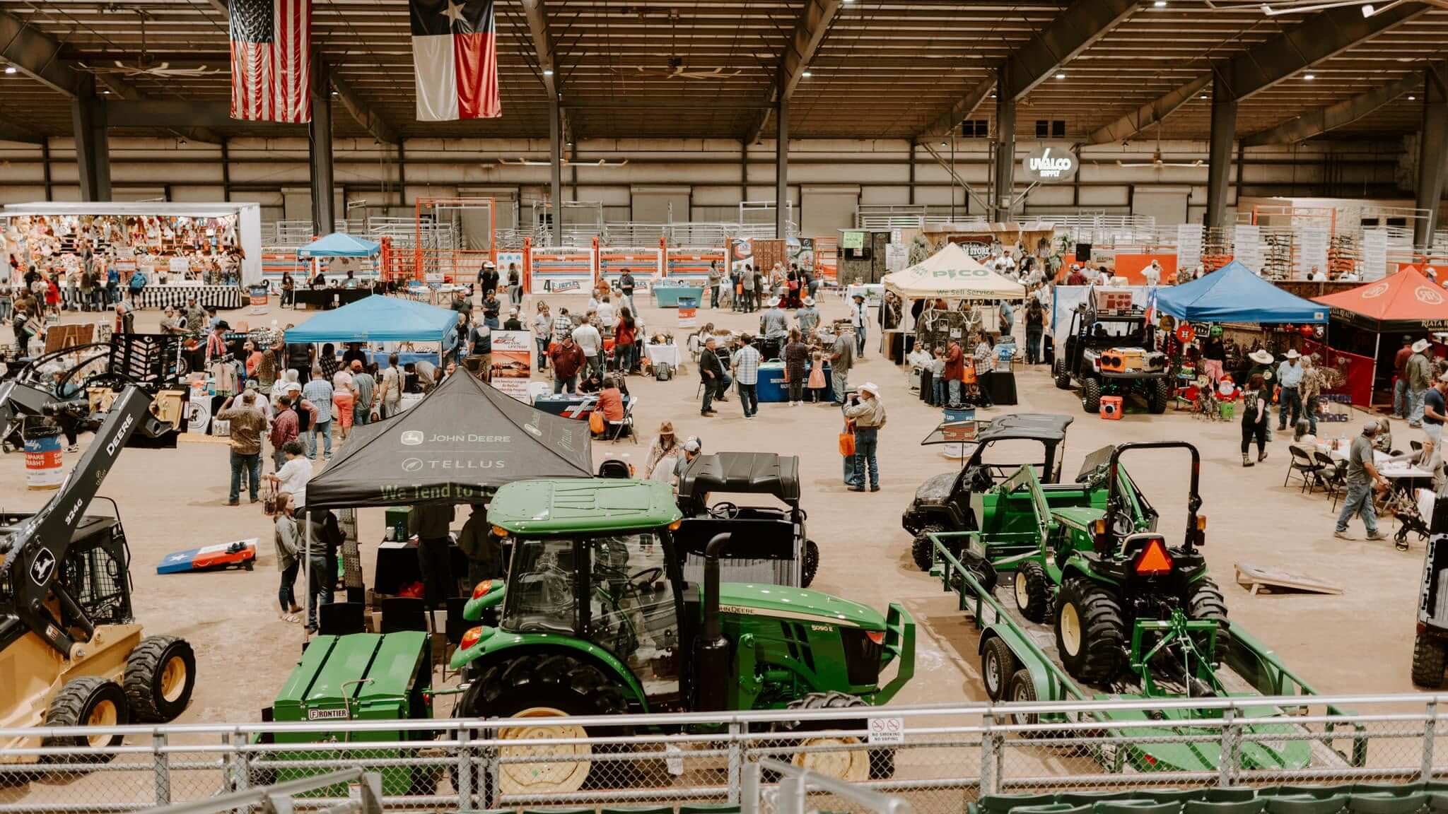 An image taken within the William.R. Mitchell Uvalde COunty Fairplex of Hunters Roundup, many booths with people standing around them are on the floor, as well as a large green tractor